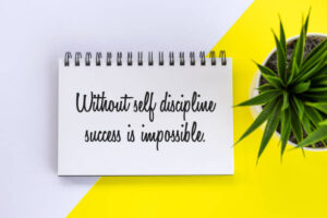 An image of the Power of Self-Discipline to Achieve Long-Term Goals with a positive motivational note on a book - Without self discipline success is impossible. Business flat lay with plant in a small pot on the white and yellow table background. Be discipline concept.