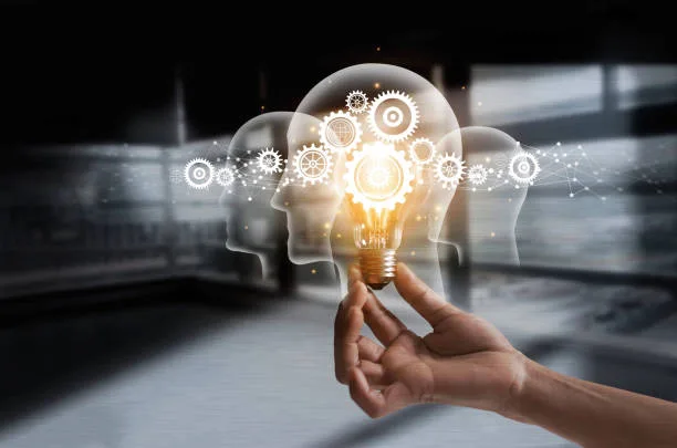 Illustration of a hand holding a glowing light bulb with overlapping human heads and gears, symbolizing creative thinking, problem-solving, and an innovation mindset.